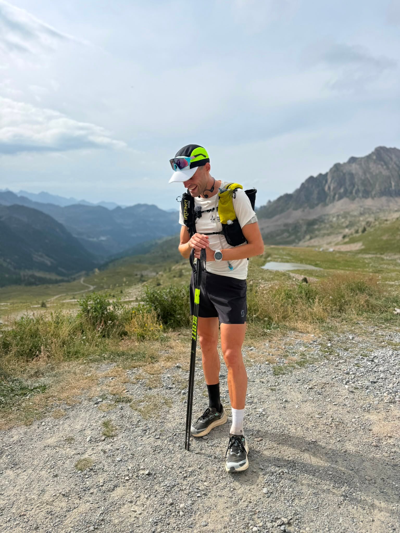 A man laughing and leaning on a trekking pole on a high pass, with a valley behind.