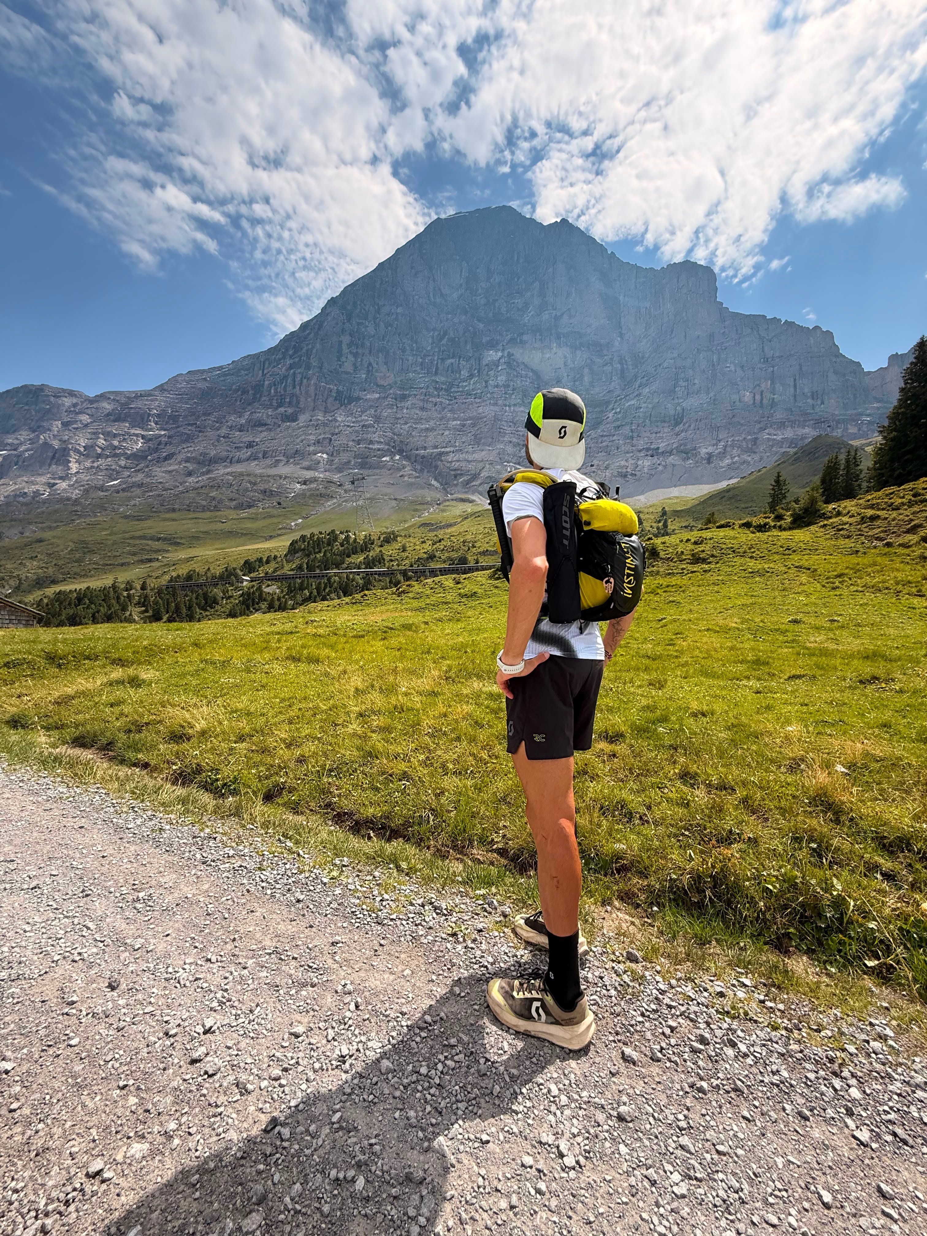 A runner seen from behind, facing a huge rock face above green meadows.