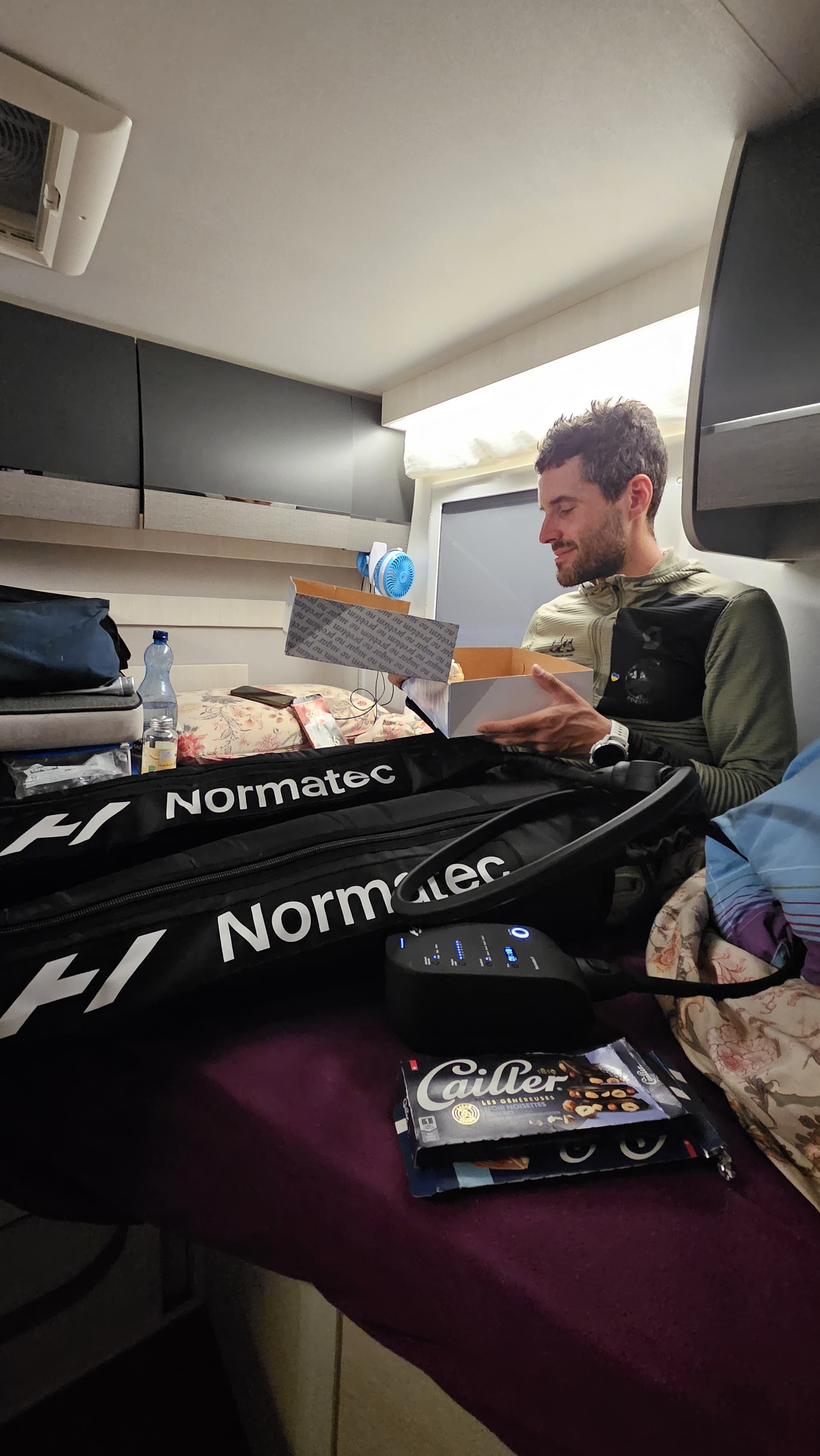 A man in a camper opening a box, with compression boots and chocolate beside him.