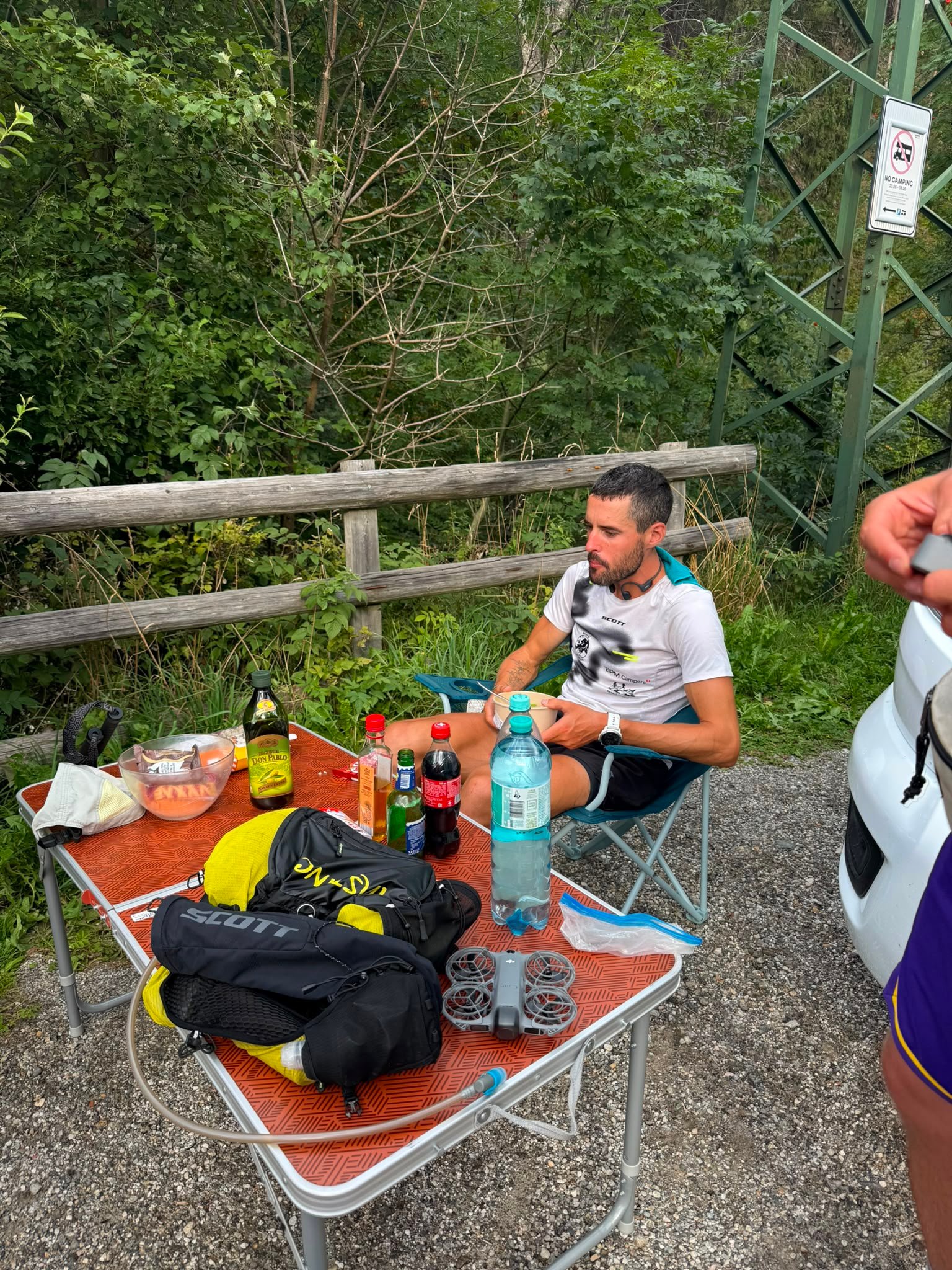 A man eating at a camp table with his gear and a drone next to him.