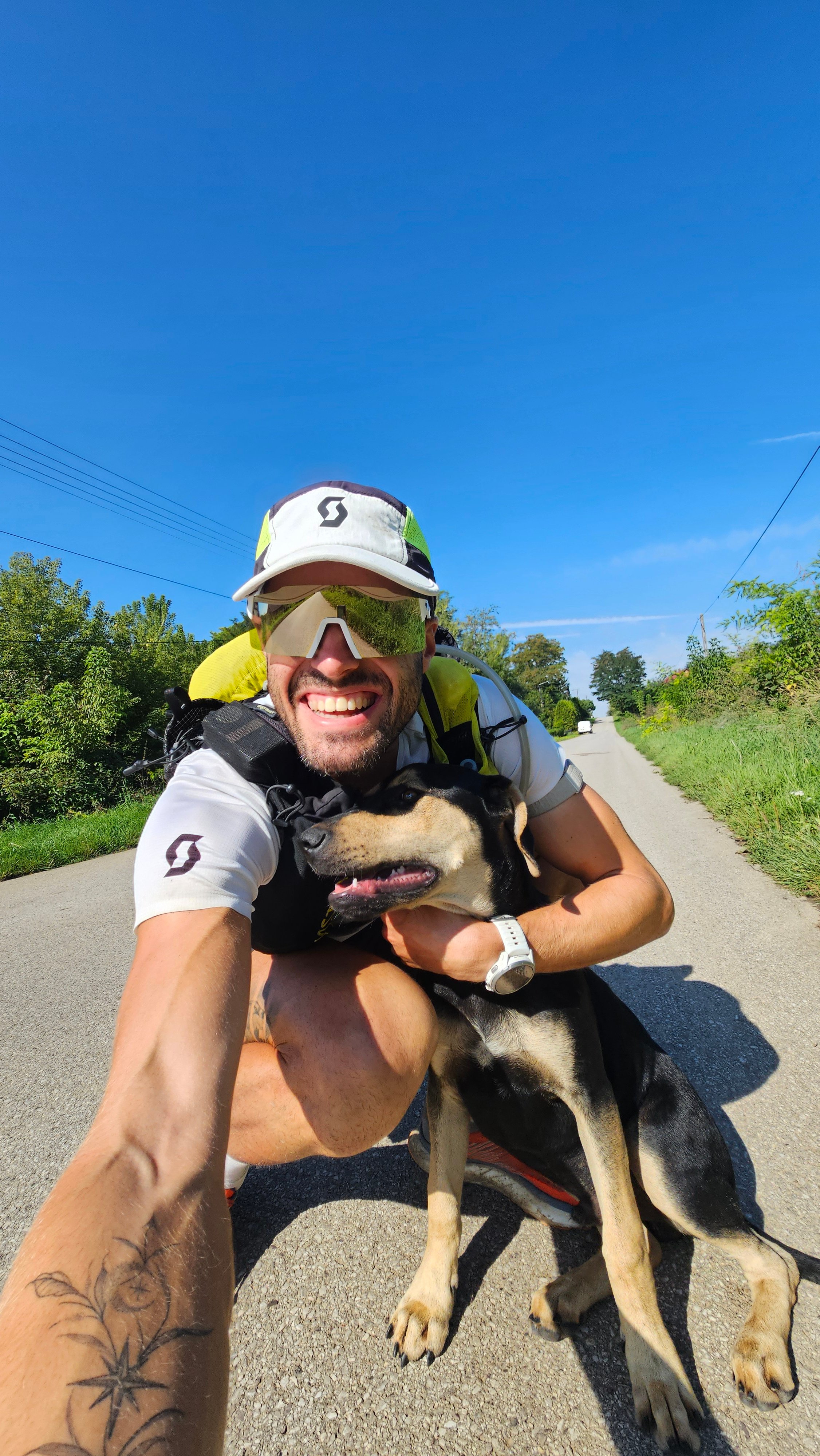 A man crouching on a rural road, hugging a dog, in a wide-angle selfie.