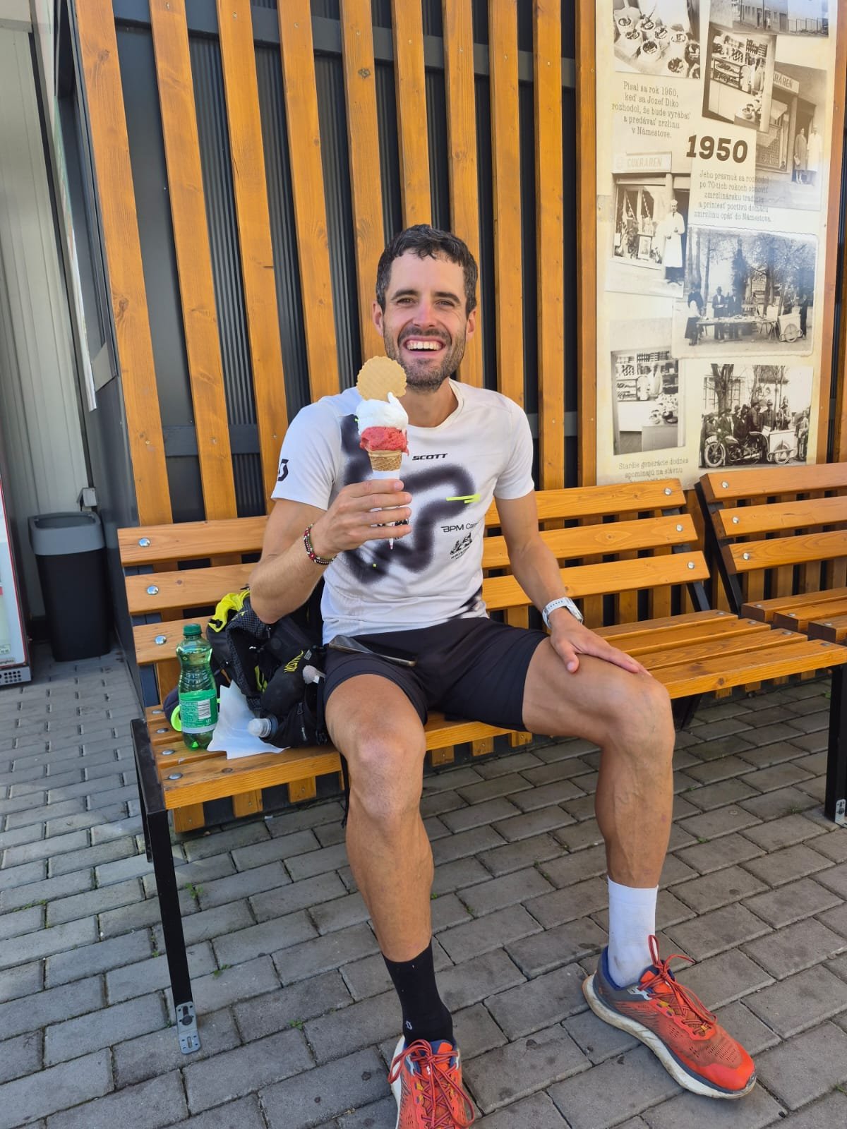 A smiling man on a bench with an ice cream cone, next to a wall panel of old photographs.