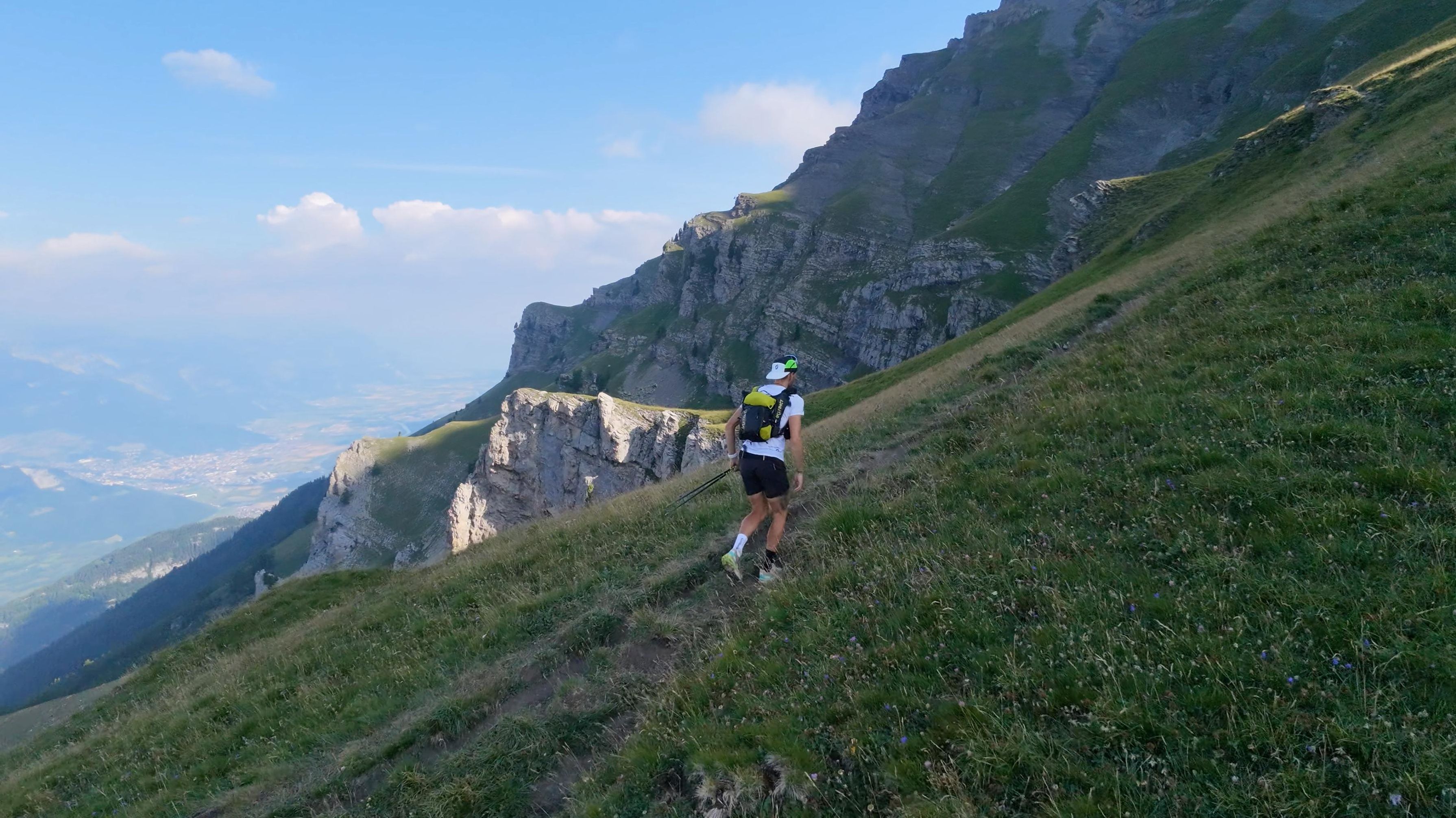 A runner seen from behind on a steep Alpine grass path above cliffs and a hazy valley.