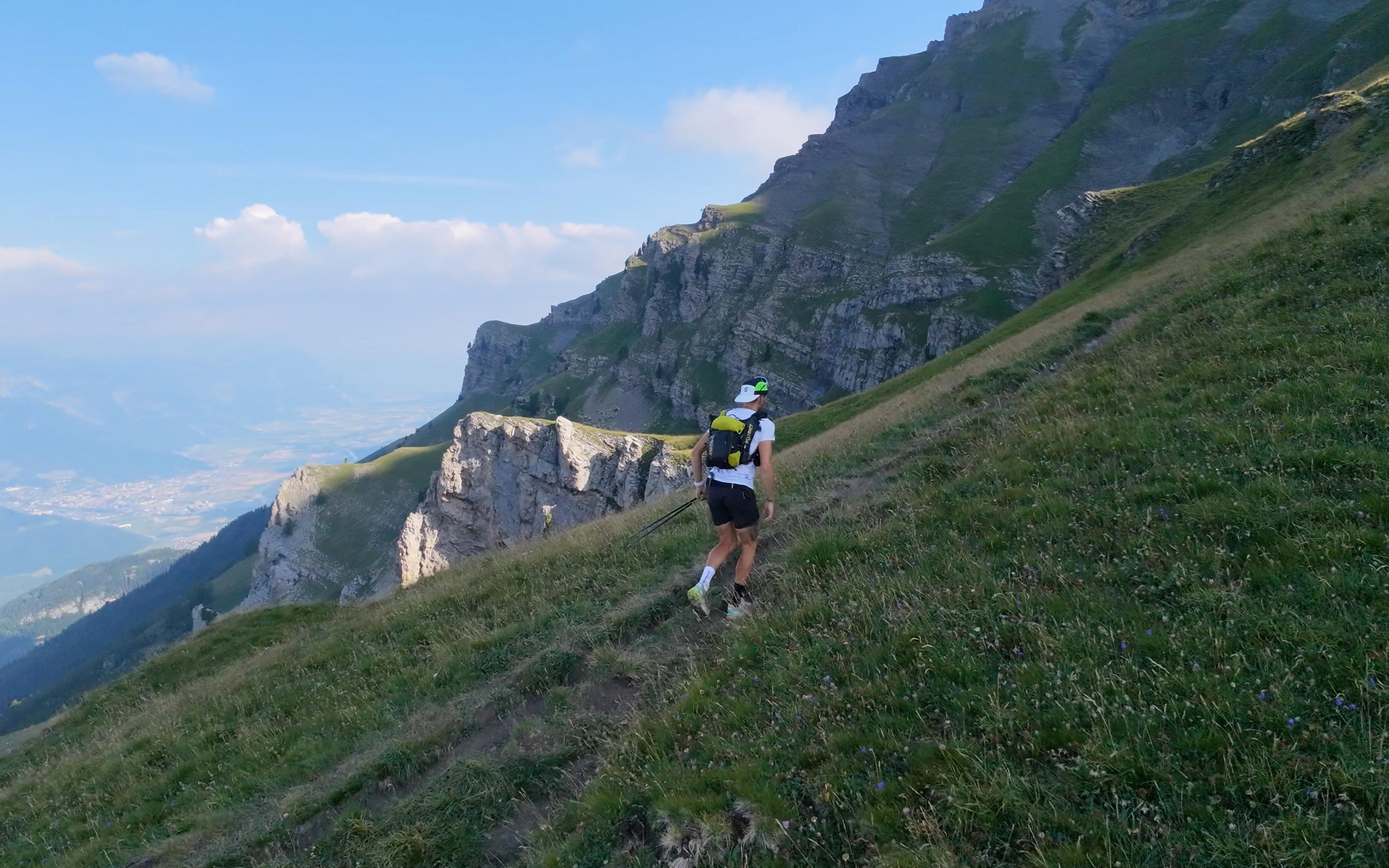 A runner seen from behind on a steep Alpine grass path above cliffs and a hazy valley.