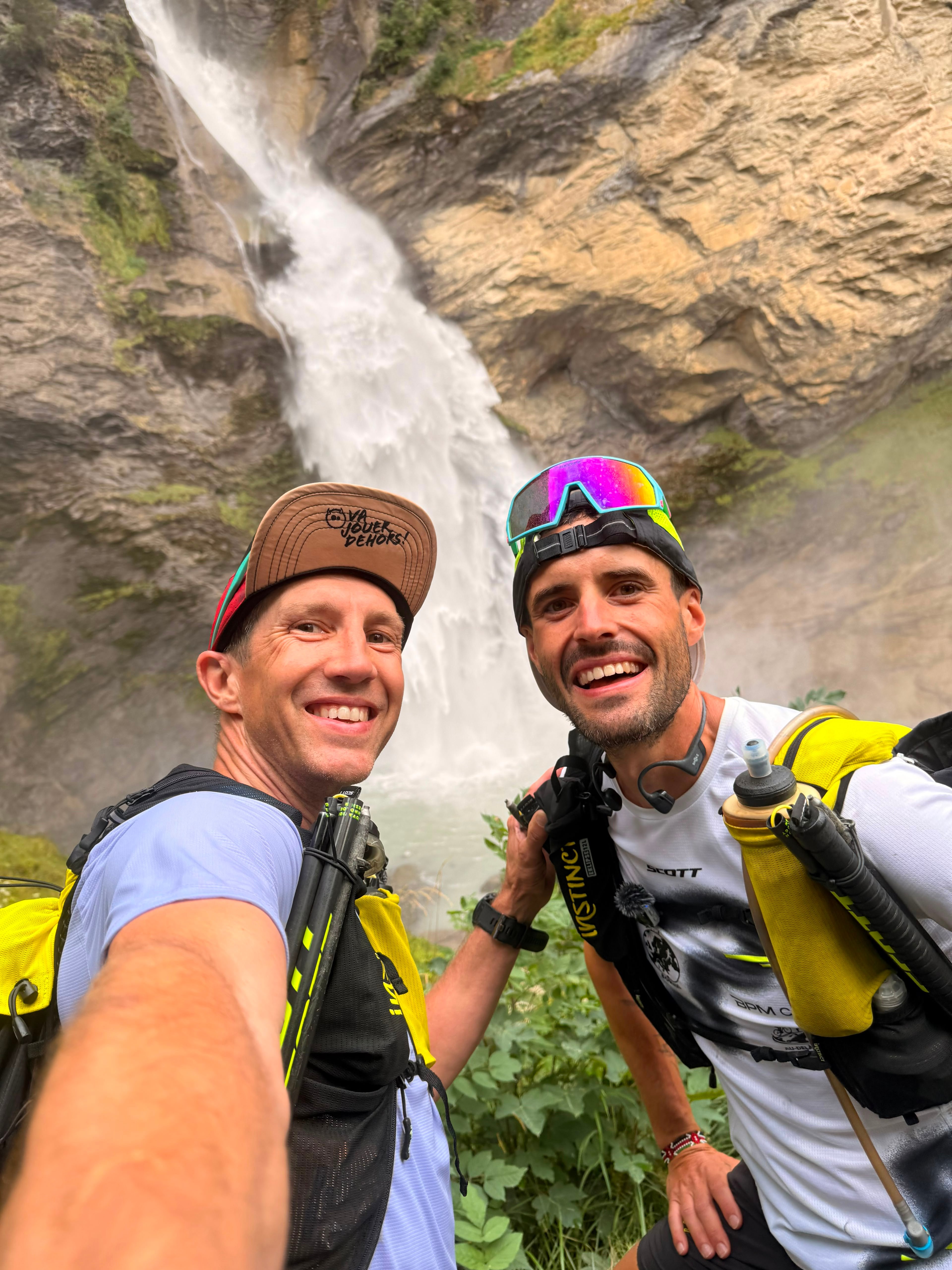 Two men in a selfie in front of a tall waterfall.