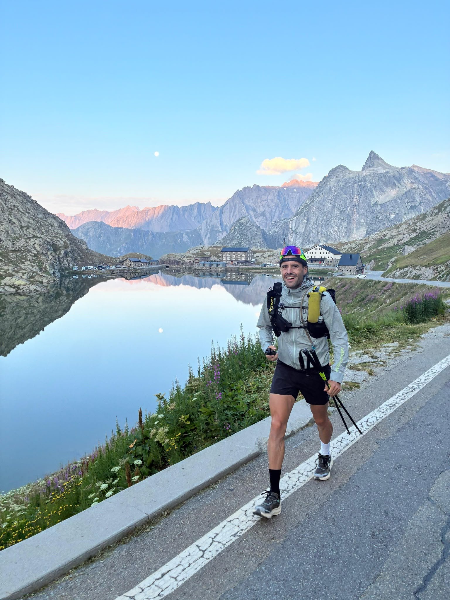 A runner on a road beside an Alpine lake at dusk, with the moon visible.