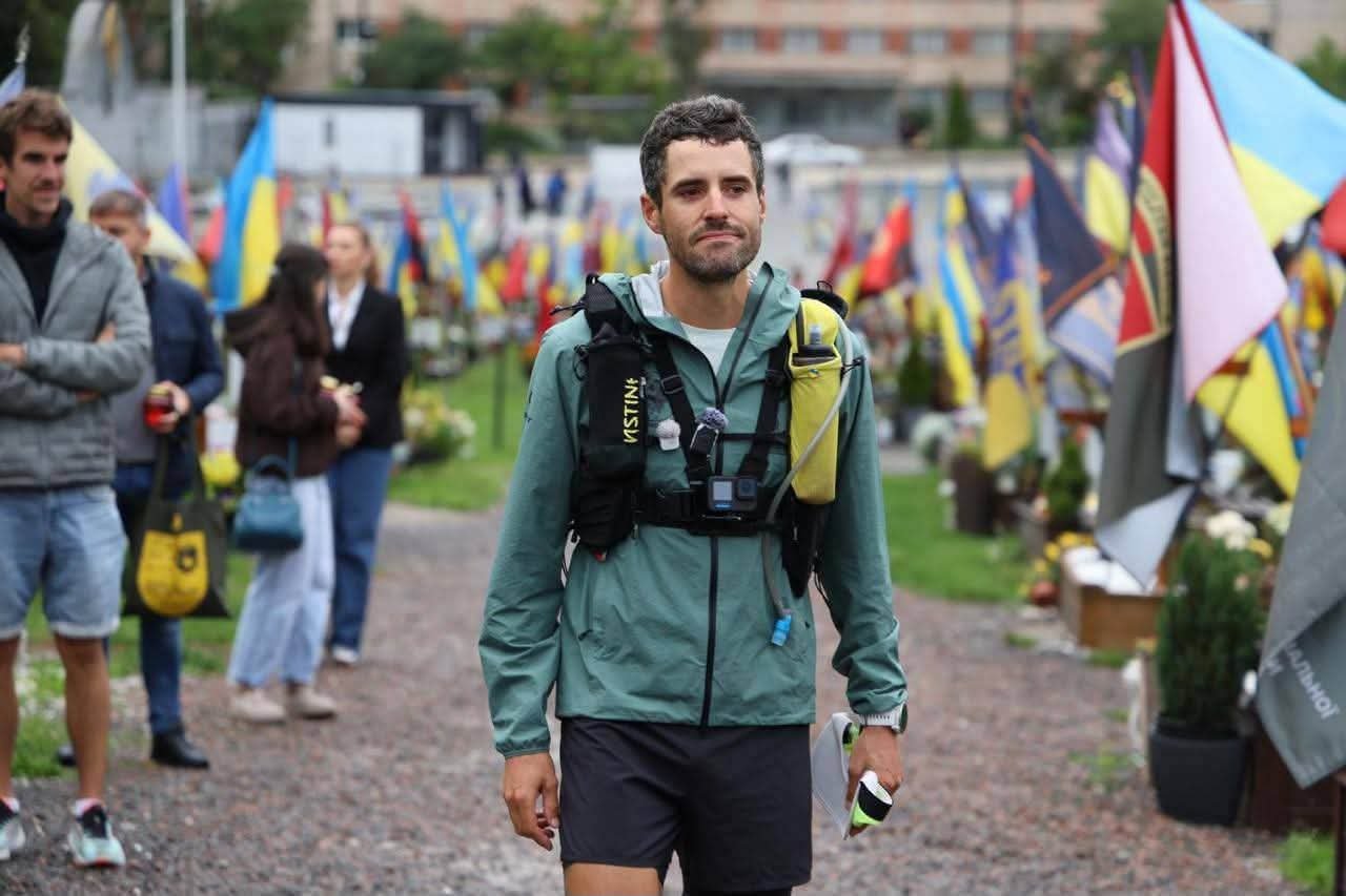 A runner in a teal jacket and running vest walking on a gravel path among flags.