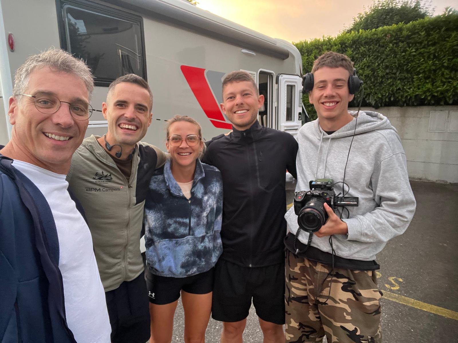Five people smiling in a selfie in front of a camper van, one holding a video camera.
