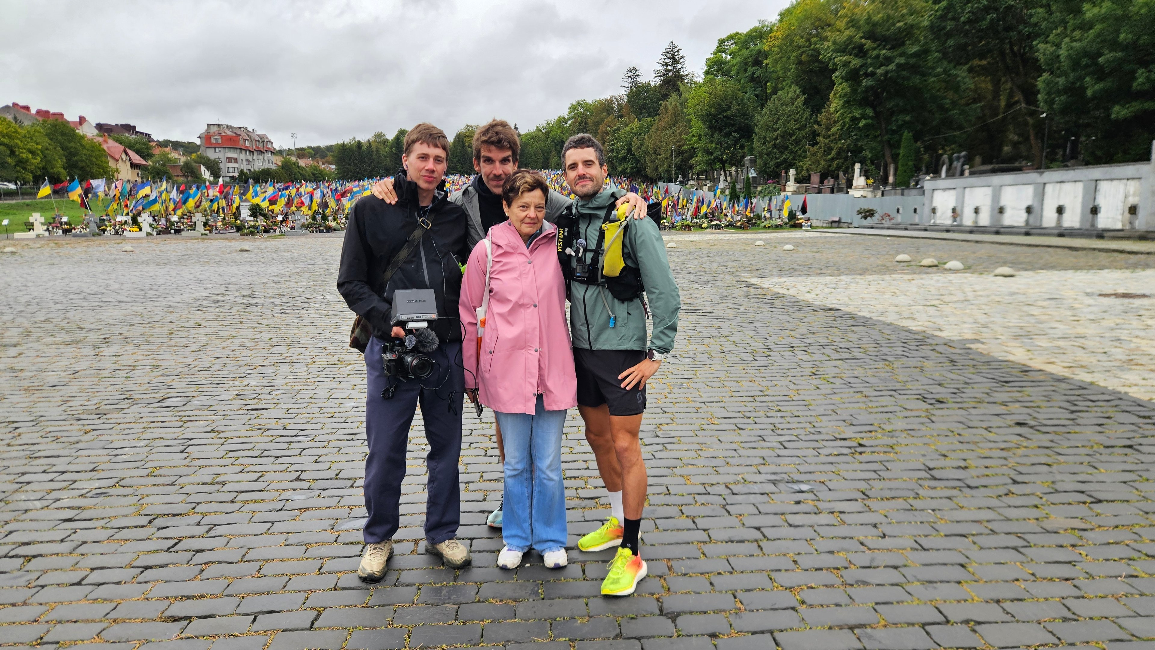 Four people posed on cobblestones with a field of blue and yellow flags behind them.