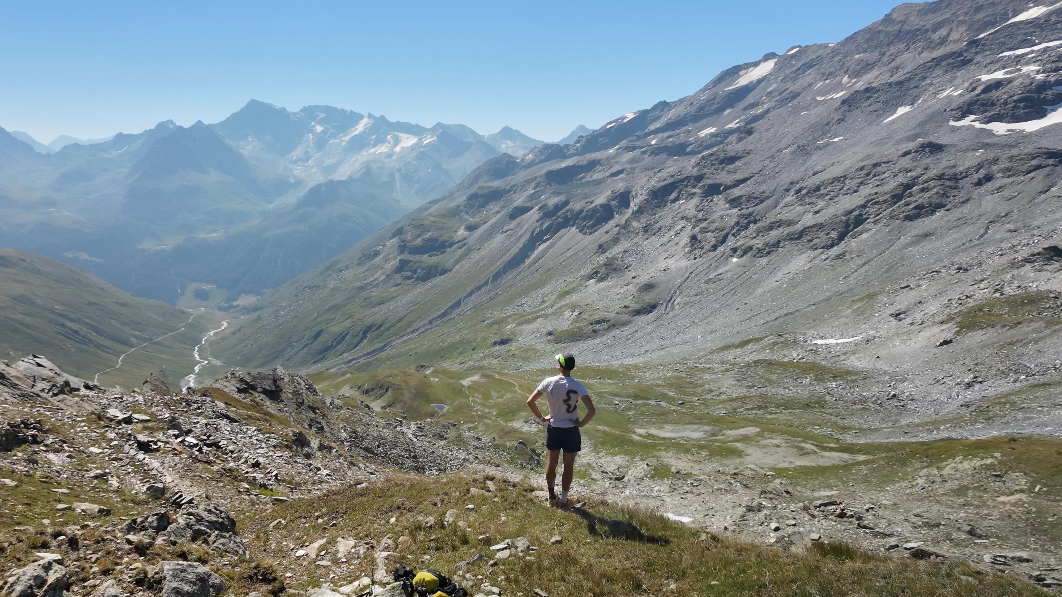 A runner seen from behind looking over a high Alpine valley with snow patches.