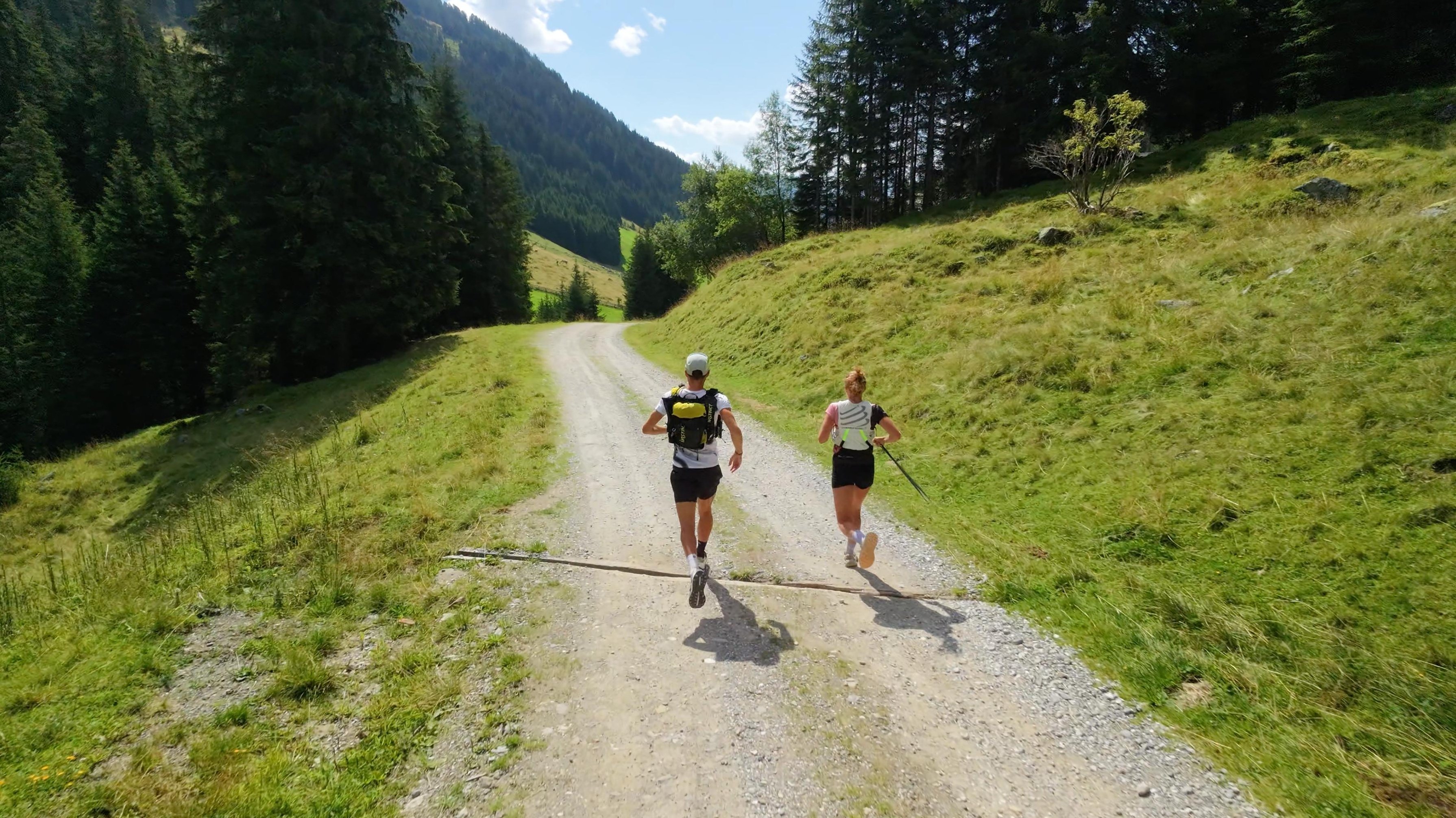 Frédéric and his girlfriend Morgane running on a gravel track through a sunny valley of conifers.
