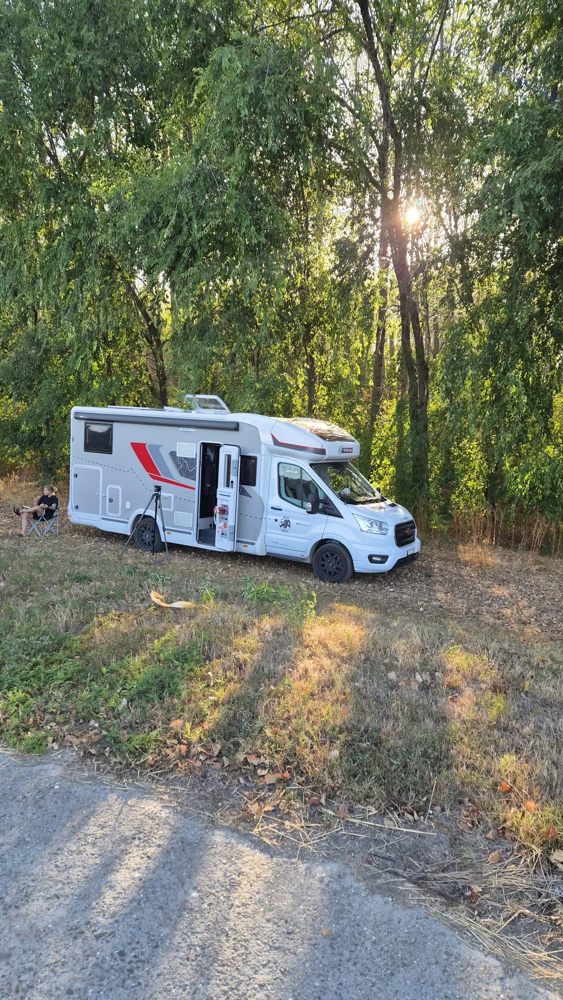A white camper van under trees with a tripod and a person sitting in a chair.