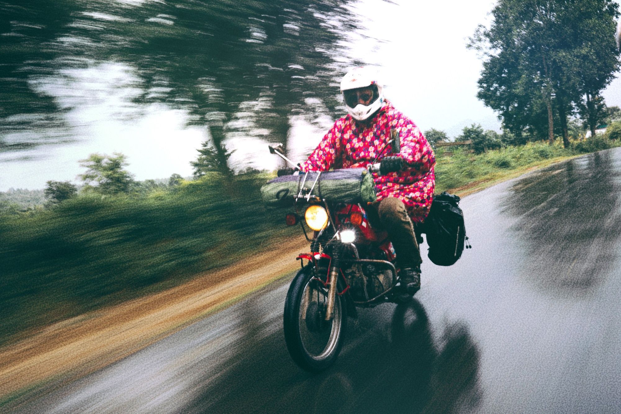 Liam Della in a pink floral rain poncho riding his loaded red Honda CT110 along a wet road, trees blurred by the camera's pan
