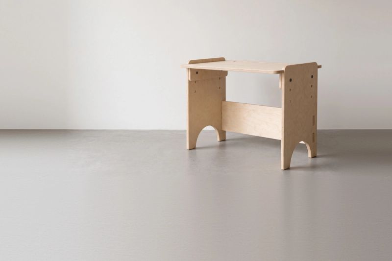 A birch plywood children's desk standing in front of a plain pale wall, with height-adjustment holes drilled in both side panels