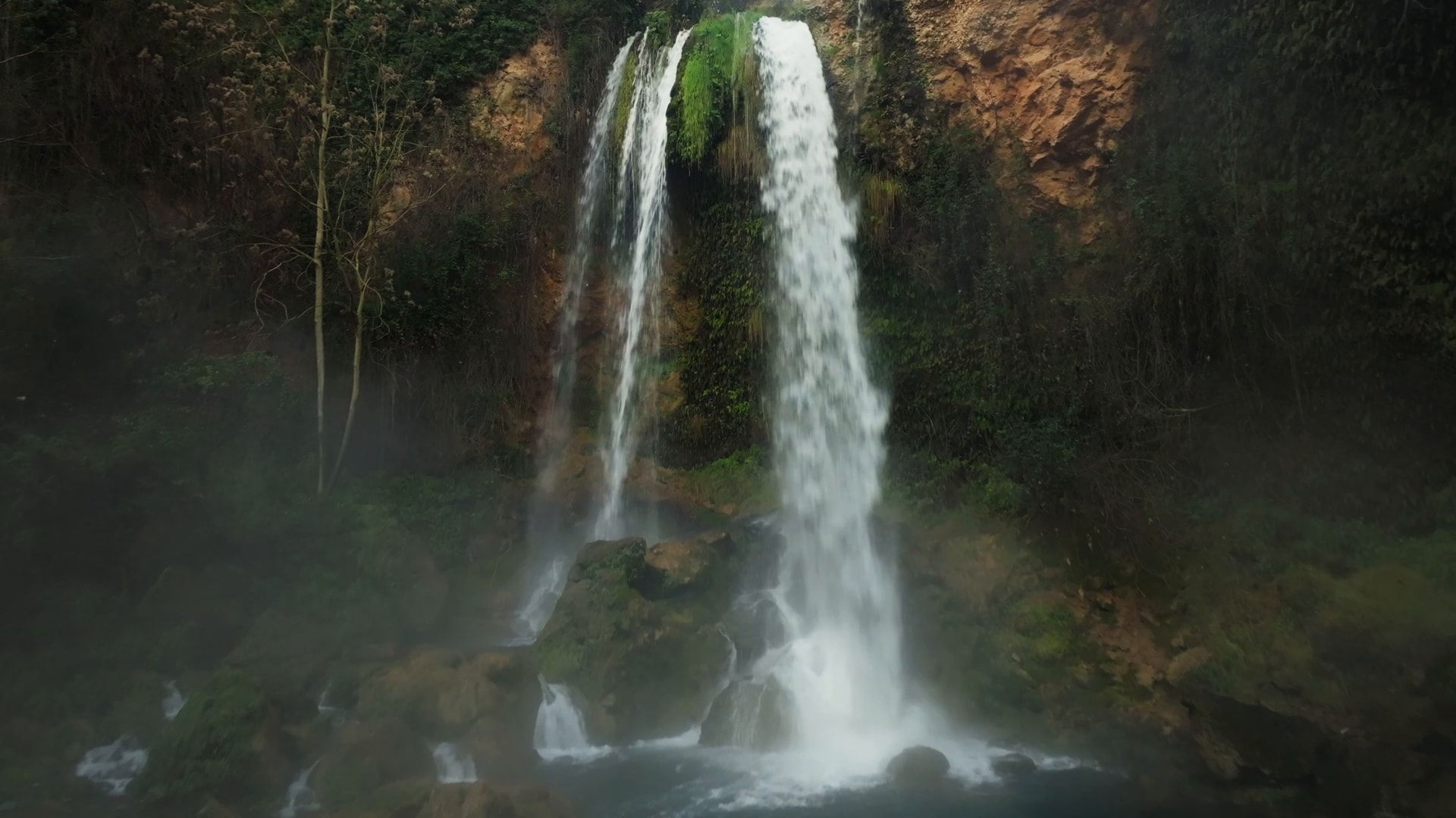 A waterfall falling through the mist into a green pool
