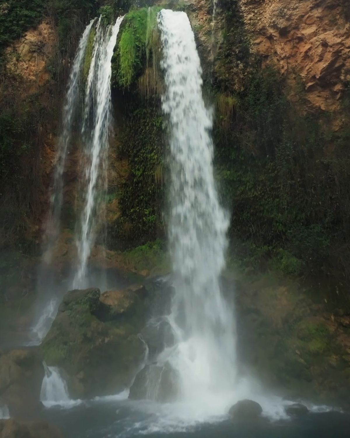 A waterfall falling through the mist into a green pool