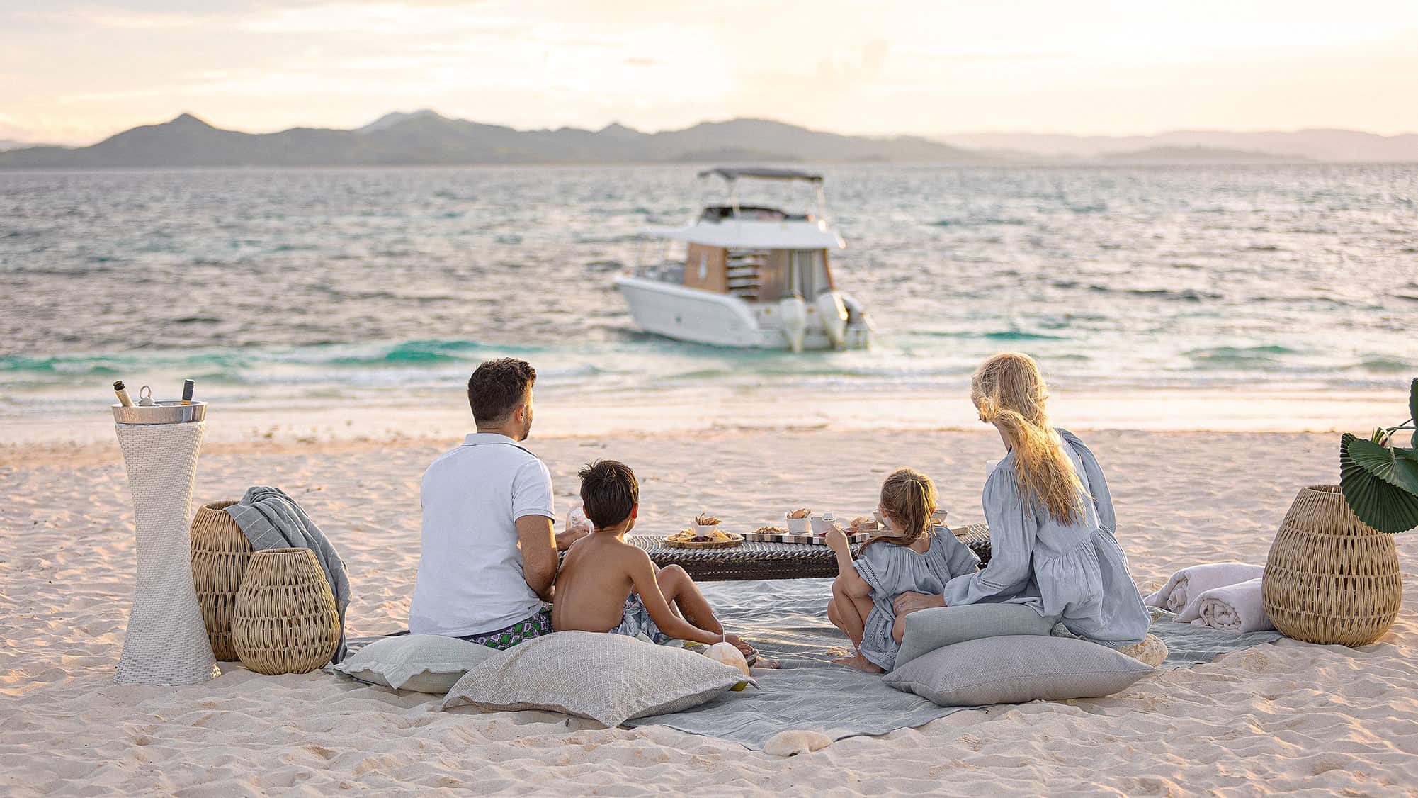 A family dining on cushions on the beach, a boat moored offshore