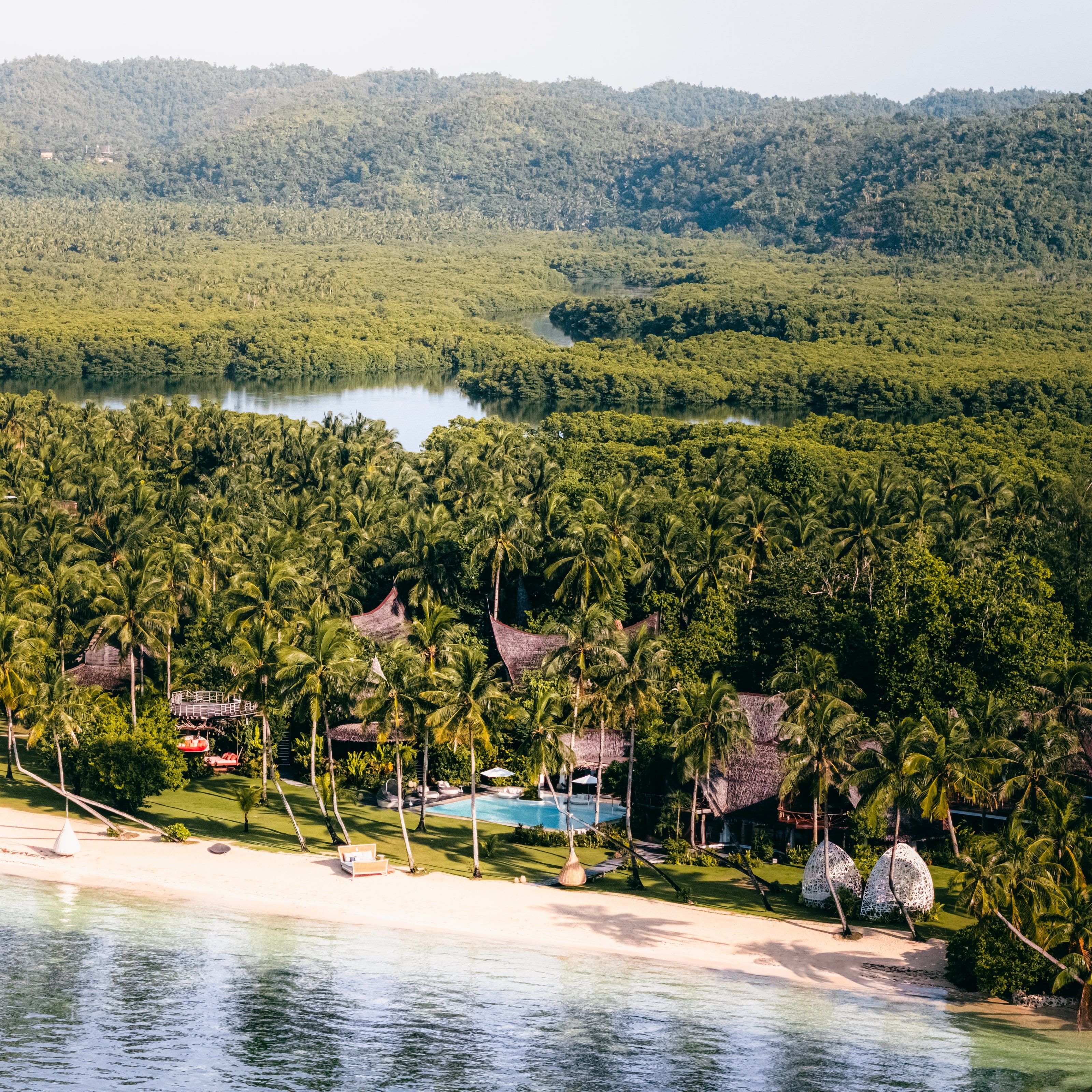 Aerial view of the resort: thatched villas and a pool among palm trees, with the mangroves behind