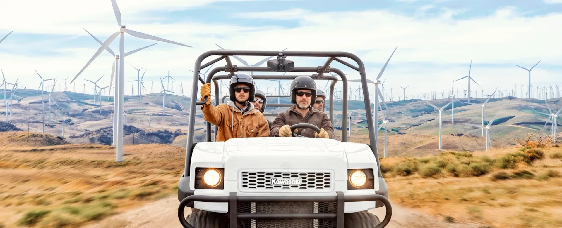 Two men in helmets riding in a Kawasaki UTV at a work site with windmills.