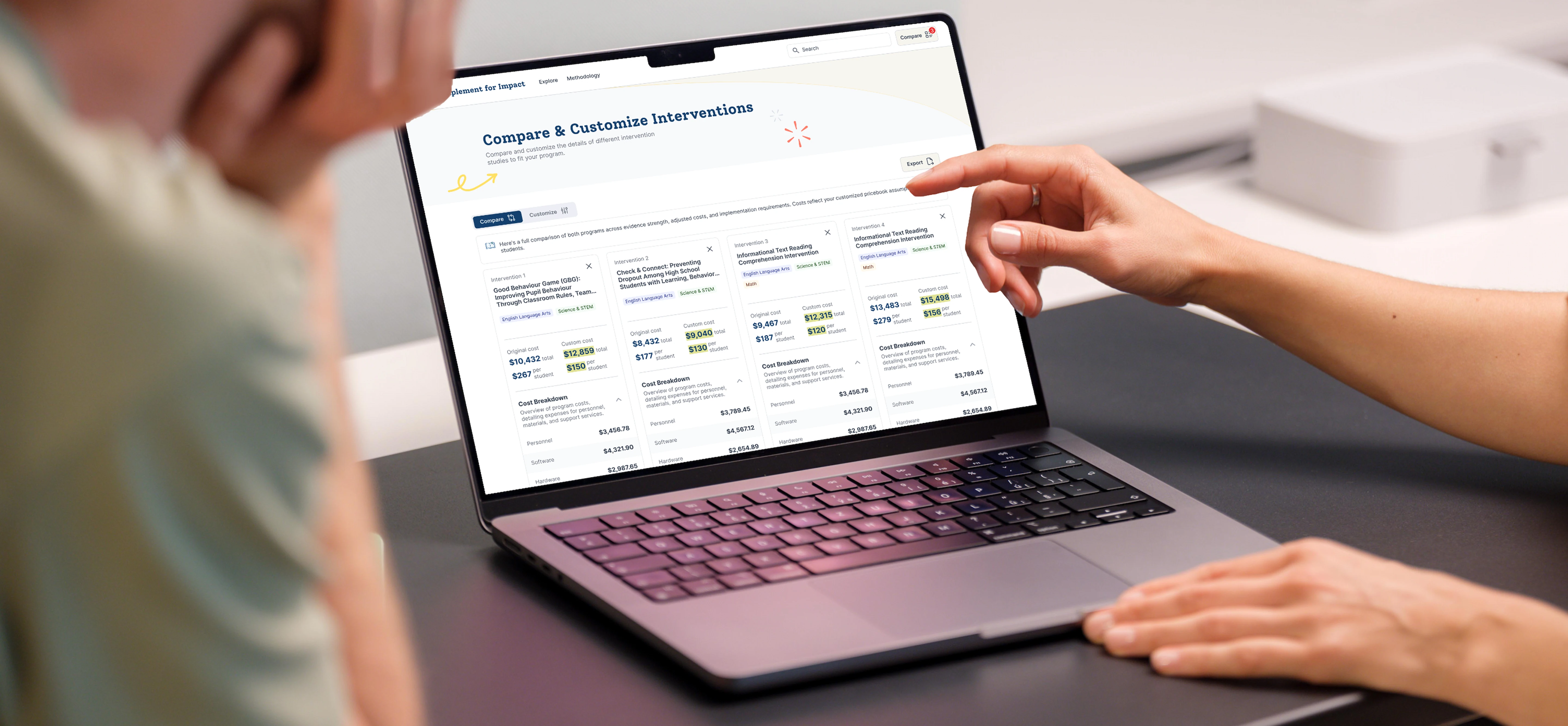 Two people reviewing the Implement for Impact website on a laptop, comparing educational interventions by cost, implementation requirements, and research findings.