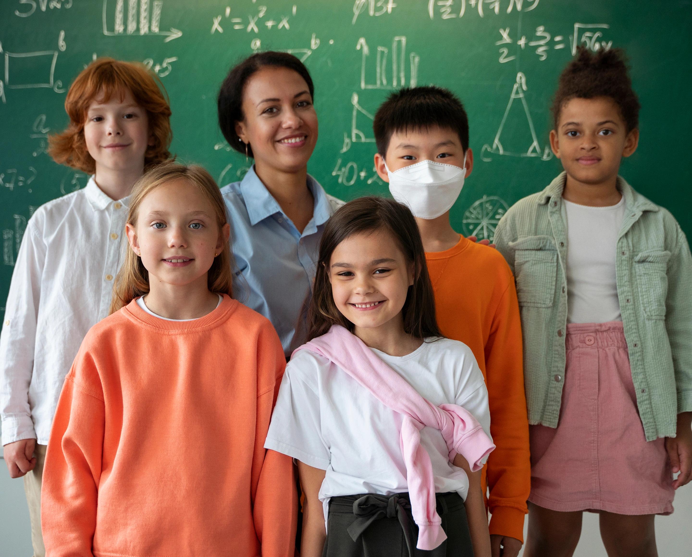 A smiling teacher with five elementary school students in front of a chalkboard covered in math equations.