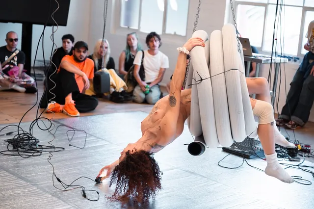 A naked woman is suspended from the roof hanging in a coiled pipe. The floor is silver and there are blurry audience members watching her from a far