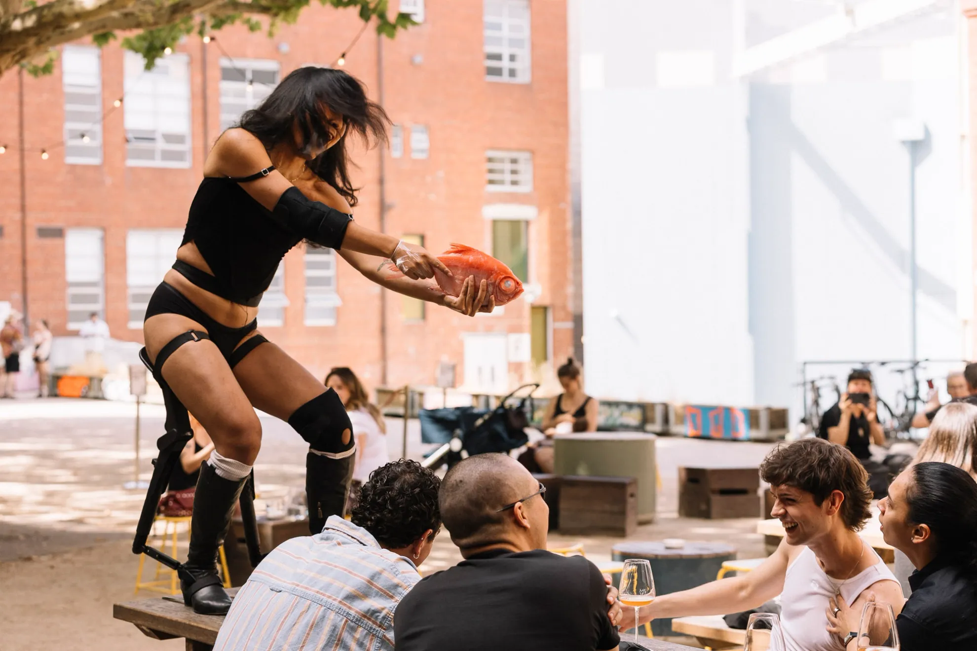 Ari standing on a table holding an orange fish like a gun pointing it at people smiling.