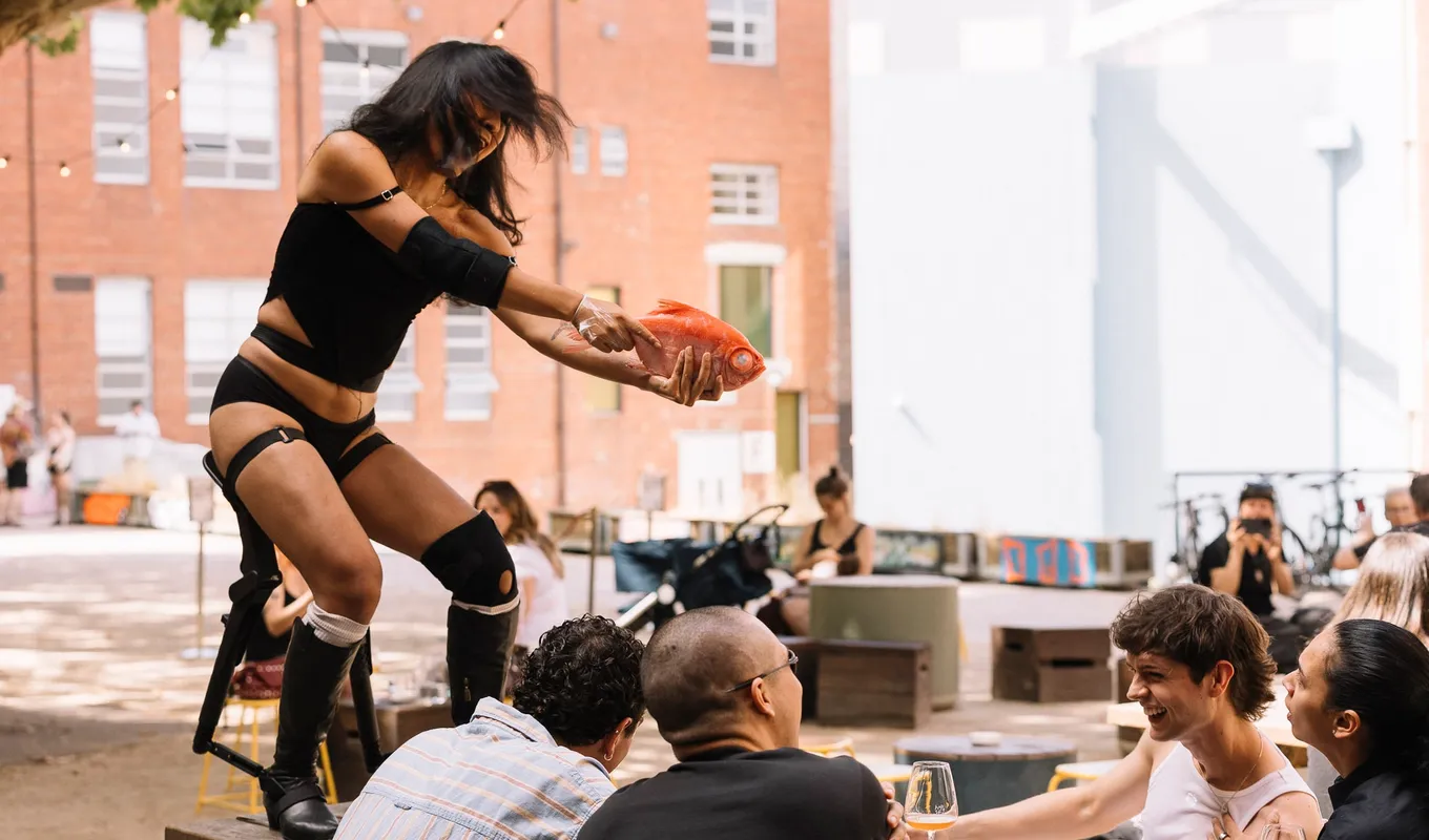 Ari standing on a table holding an orange fish like a gun pointing it at people smiling.