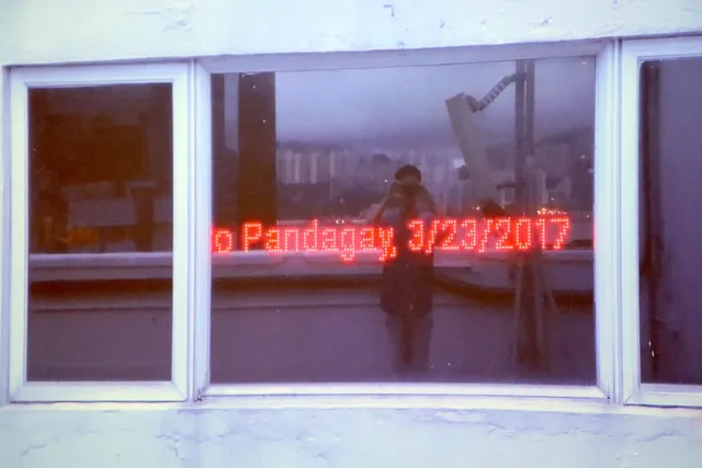 Red, glowing text that reads 'Pandagay, 3/23/207' displayed behind a short, landscape white window frame. The figure taking the photograph is visible in the reflection holding their camera. The text is digitally produced.