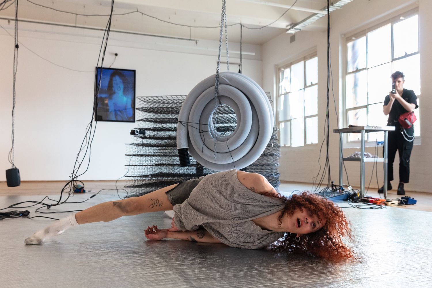 A woman lying on the foil mat on the floor. A photographer, a coil of plastic pipe that is suspended from the ceiling with chains, a pile of springs from mattresses, and a photo of Andrea are in the background.