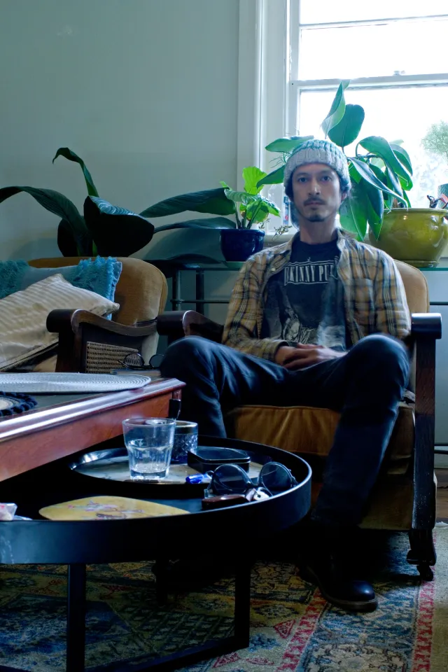 A portrait of a figure sitting in their loungeroom. They are sitting on an armchair with a table infront of them that has assorted objects on it. The figure is wearing a flannel shirt and a woolen beanie