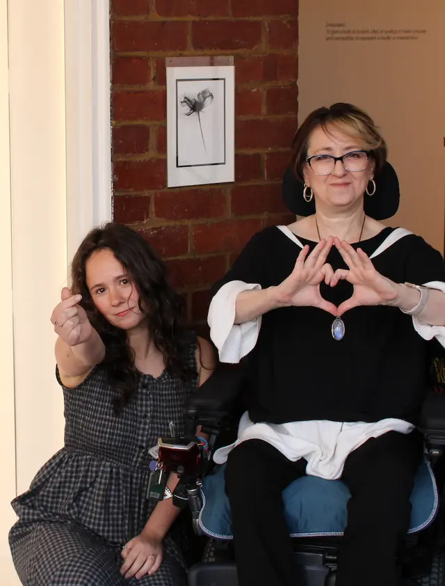 Two women are making heart gestures, one of them is on a wheelchair. A piece of white paper with a black flower on it is attached to the brick wall in the background.
