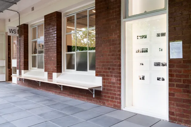 An outside view on the balcony of west space. There are many small black and white photographs in the west space window gallery. There is a sign outside the doors of the gallery on the wall with the letters WS