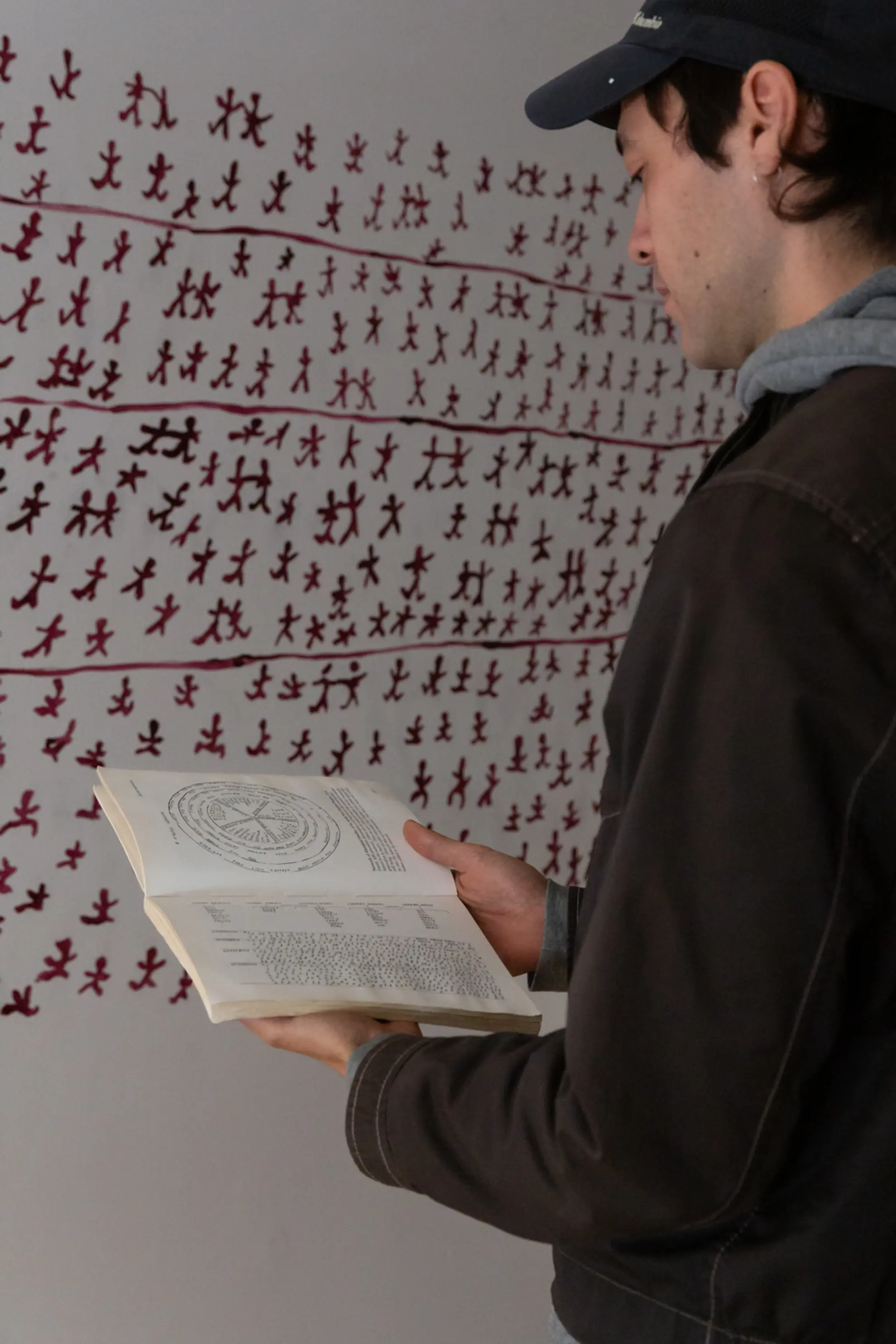 Sebastian Henry-Jones with Patsy Cohen's 1990 book 'Ingelbah and the Five Black Matriarchs'. He is standing in front of wall painting by Gabi Briggs based on original drawings by Cohen. Photography by Janelle Low.