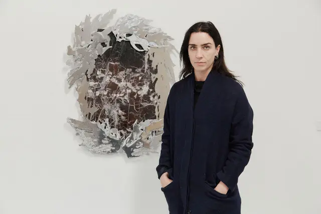 a woman with dark hair and hazel eyes stands in front of her artwork made of metal and marble displayed on a white wall. She is wearing a long dark coat with her hands in her pockets