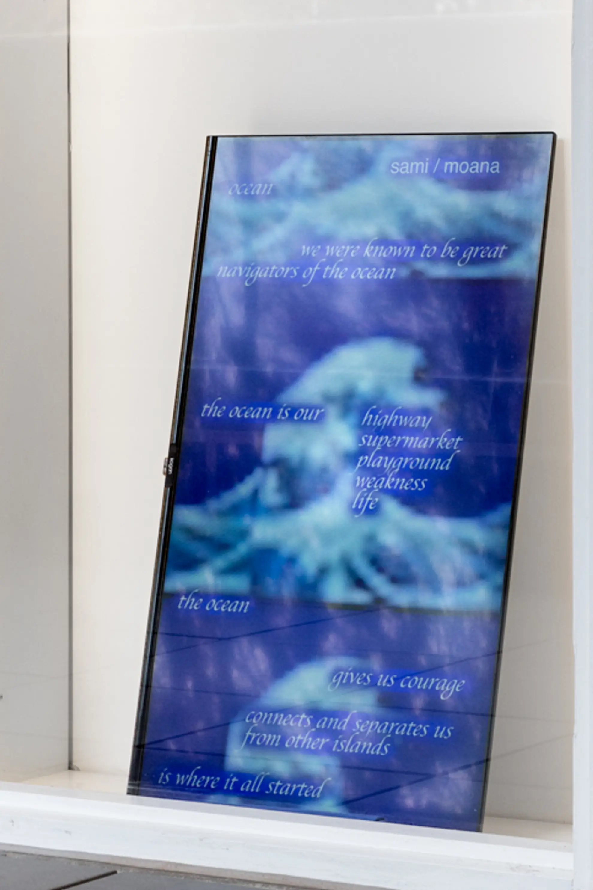 A screen leaning behind a shallow window display, on its side. The screen depicts a deep blue landscape with the silhouette of a tree in the bottom corner. There are shades of clowd.