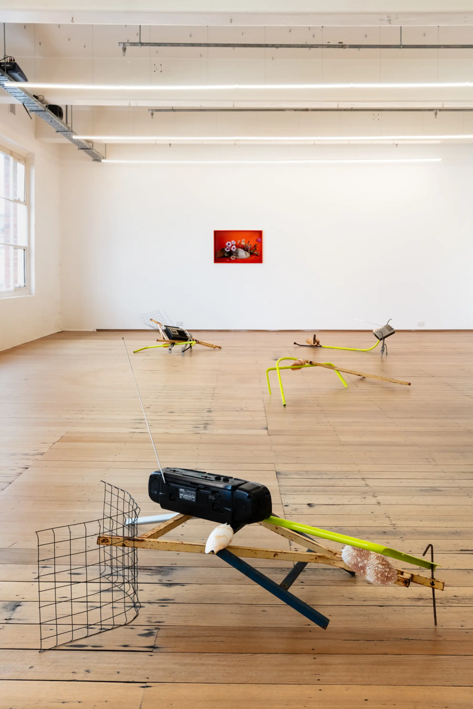 A shot of the four sculptural installation pieces sits on the gallery floor. They are comprised of metal, radios, wire frame and shells. In the background is a framed photography piece of flowers resting on another shell.