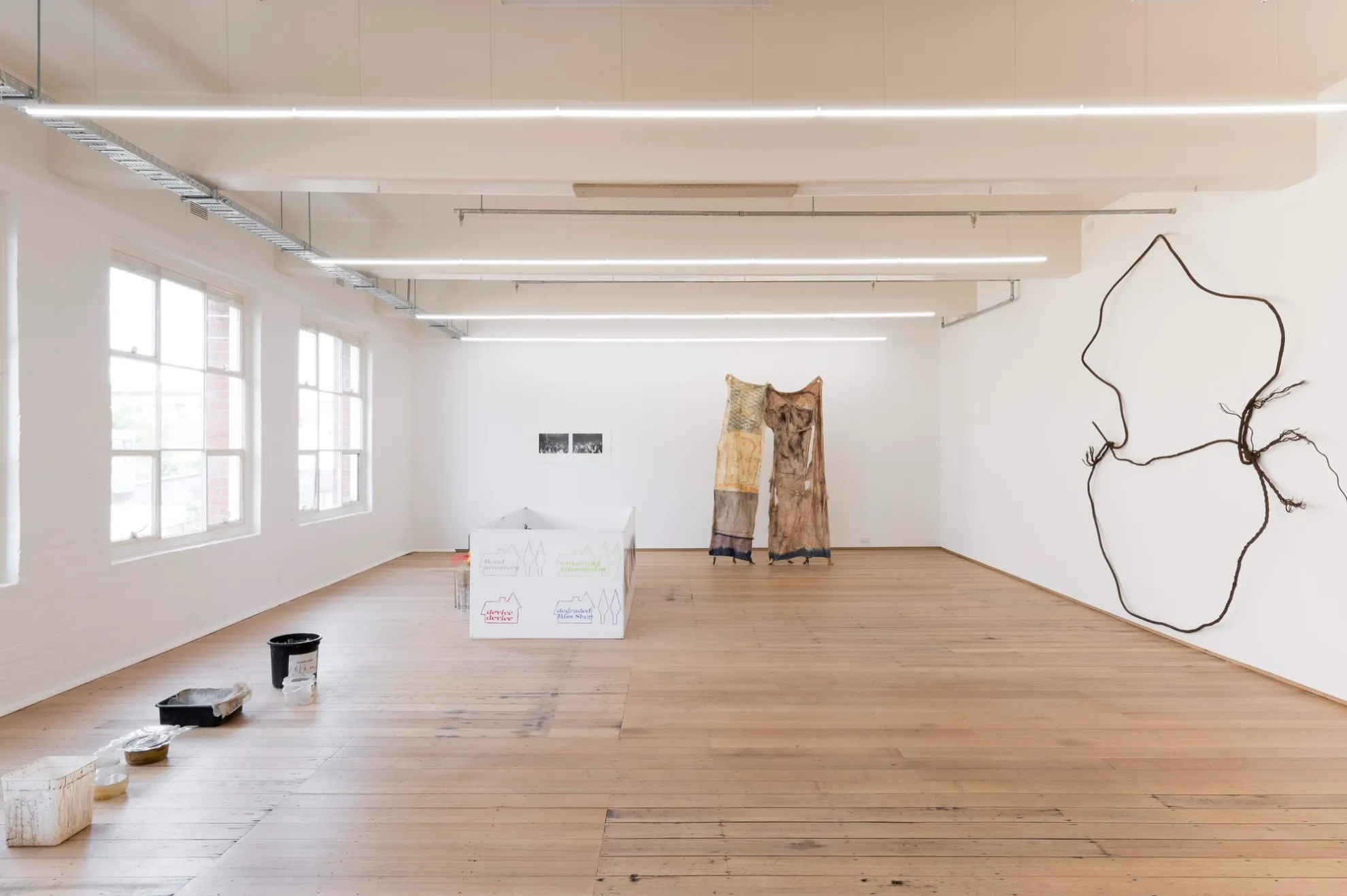 A large room with floorboards and white walls. Large industrial windows line the left hand side. 6 plastic containers of different sizes are lined up on the floor parallel to the left hand wall of the room. Going around the room clockwise, there is a large white rectangular box with colourful writing and drawings; a pair of black and white photographs on the wall; a large fabric installation in the shape of a pair of pants; and a large sculpture installed on the right-hand wall, made of rusted cable.