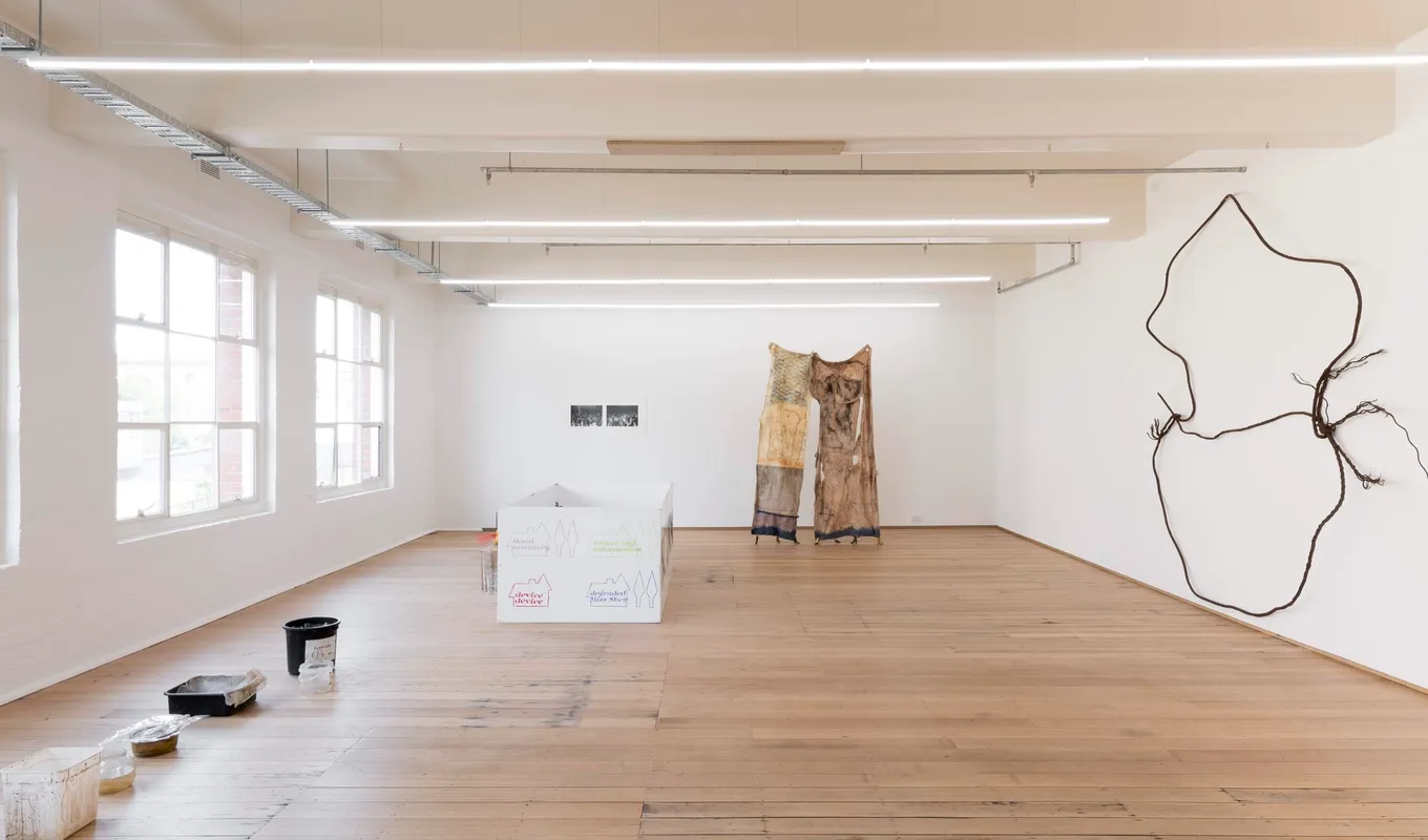 A large room with floorboards and white walls. Large industrial windows line the left hand side. 6 plastic containers of different sizes are lined up on the floor parallel to the left hand wall of the room. Going around the room clockwise, there is a large white rectangular box with colourful writing and drawings; a pair of black and white photographs on the wall; a large fabric installation in the shape of a pair of pants; and a large sculpture installed on the right-hand wall, made of rusted cable.