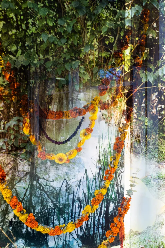 Print of green leaves, trees and water. Four strings of orange and yellow flowers hung across the frame, one beaded string.