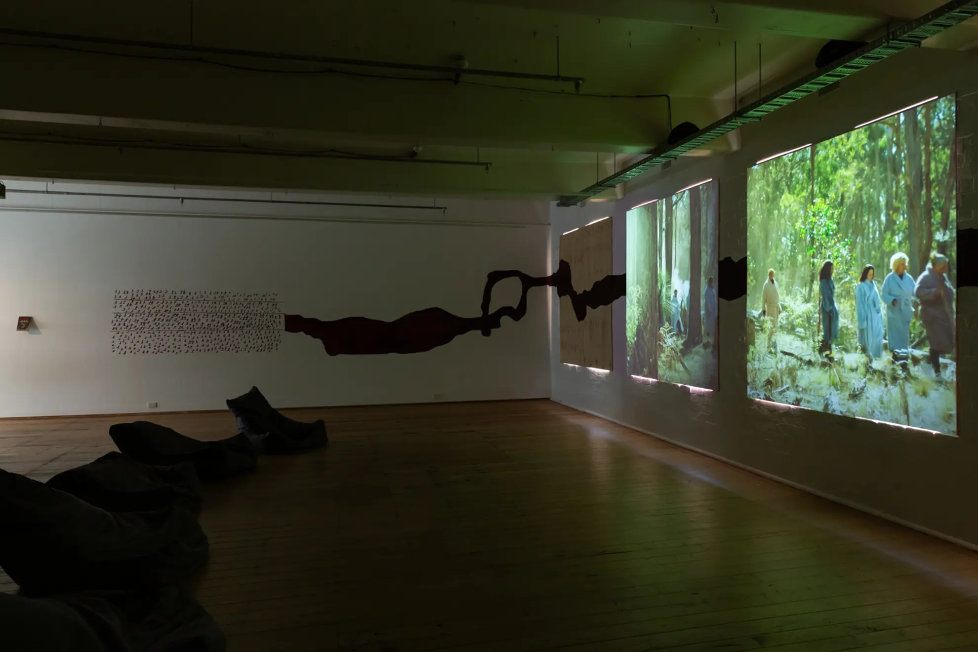 An art installation featuring three large projection screens displaying scenes of people in the bush, with a shadowy black silhouette running horizontally across the wall. The room has wooden flooring and several black bean bags positioned in front of the projections.