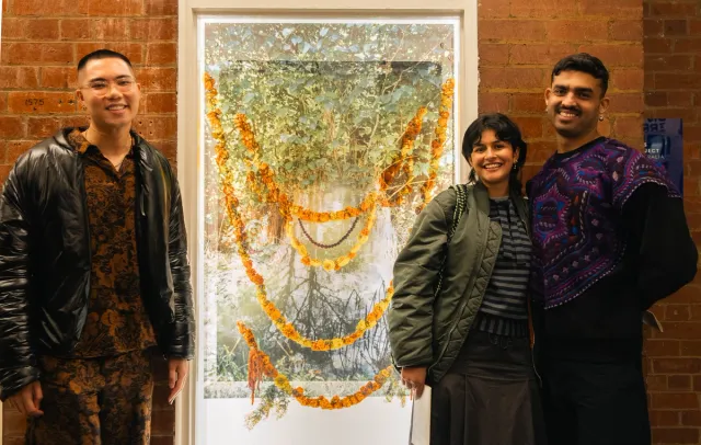 Three people posing for a photo in front of a brick wall and window display. Large bright window display with photo prints of trees, beads and orange and yellow flowers hung across the frame.