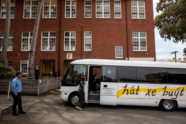 White bus parked on the road in front of a brick building with eighteen windows and trees surrounding. The bus door is open with one person is sitting inside, a woman is leaning on the bus and a man is standing on the left looking at the bus.