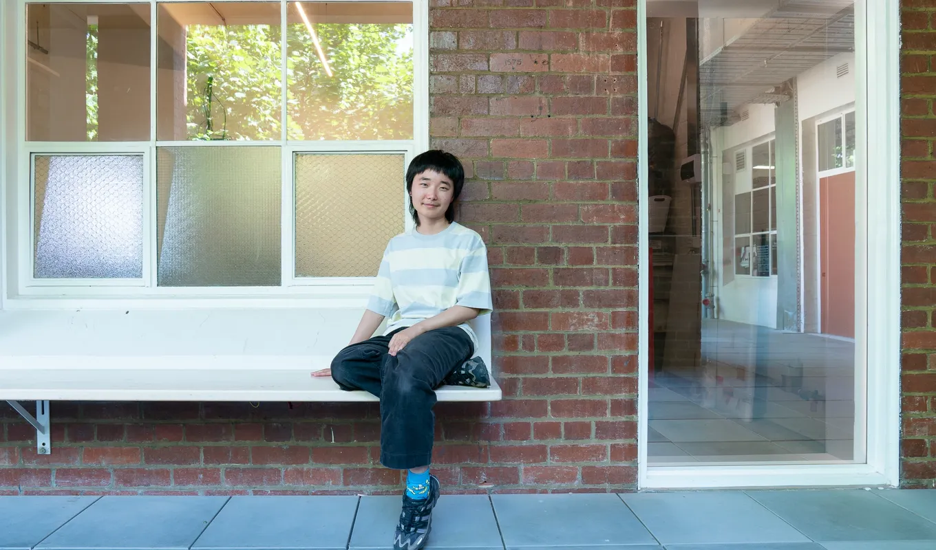 Artist Joy Zhou sits on a bench outside West Space, next to the West Space Window, which they have turned into a see-through mirror, reflecting both outside the Window and making what is inside visible - such as a tool trolley and a ladder.