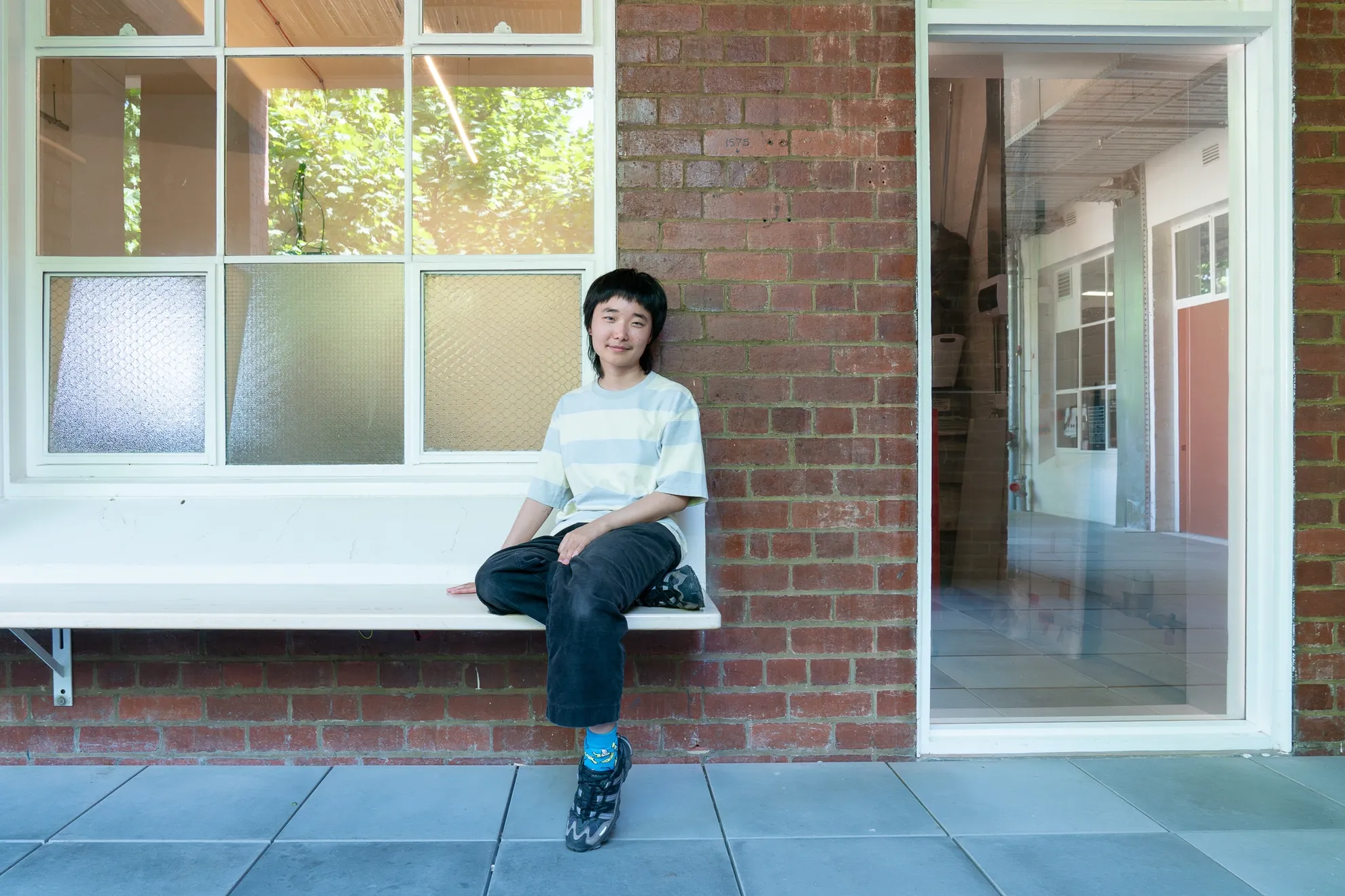Artist Joy Zhou sits on a bench outside West Space, next to the West Space Window, which they have turned into a see-through mirror, reflecting both outside the Window and making what is inside visible - such as a tool trolley and a ladder.