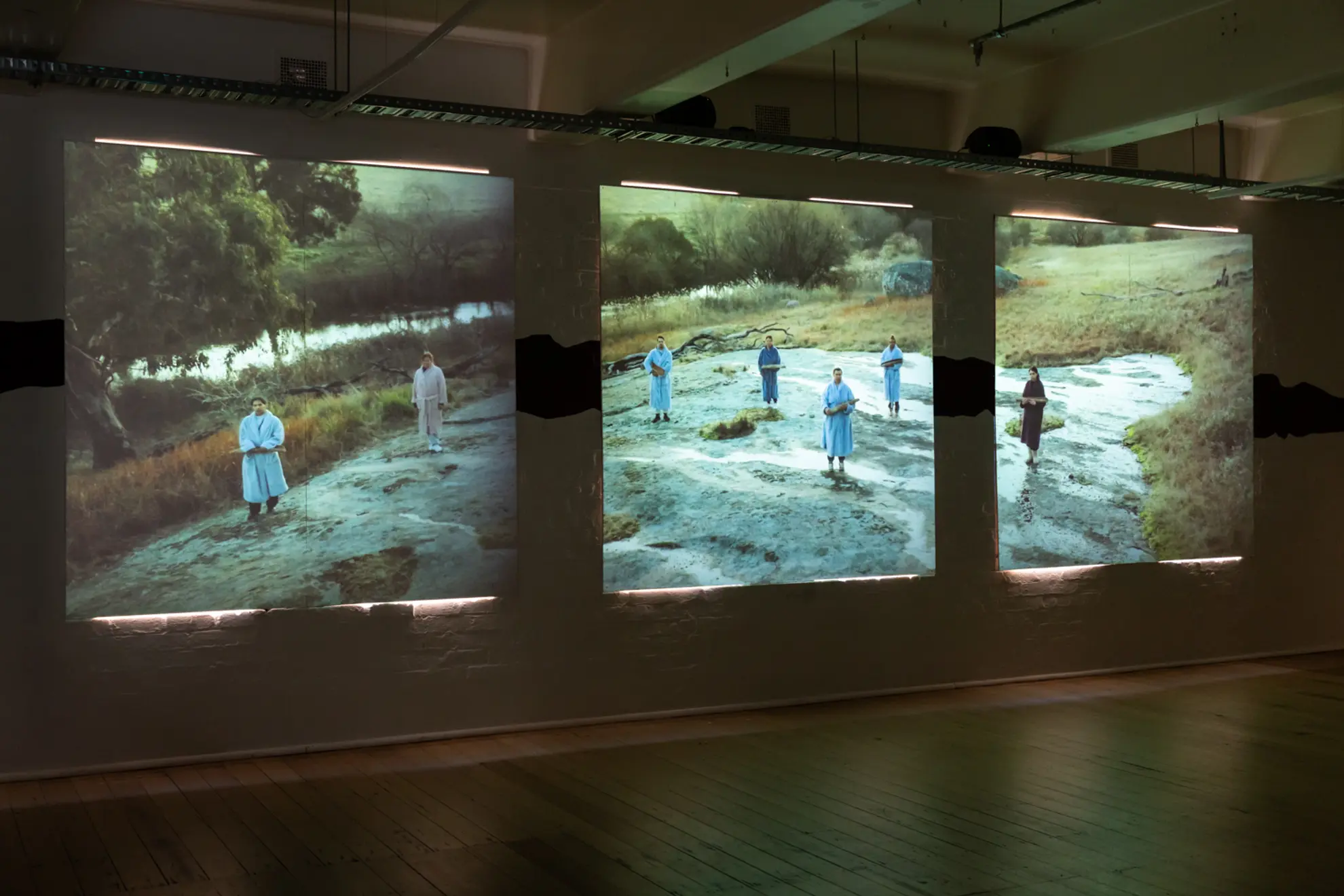 Three square video panels projected onto a wall showing seven people standing straight on, holding wooden objects.