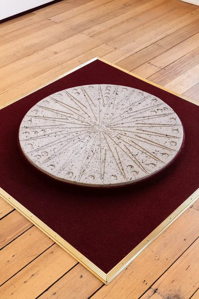 a large circular grey disc sits on a large square of burgundy carpet on top of floor boards.