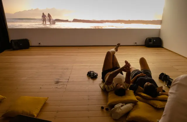 2 figures lay on the ground at West Space with yellow pillows under their heads. They are watching a projection of two female surfers walking with their boards out to the ocean