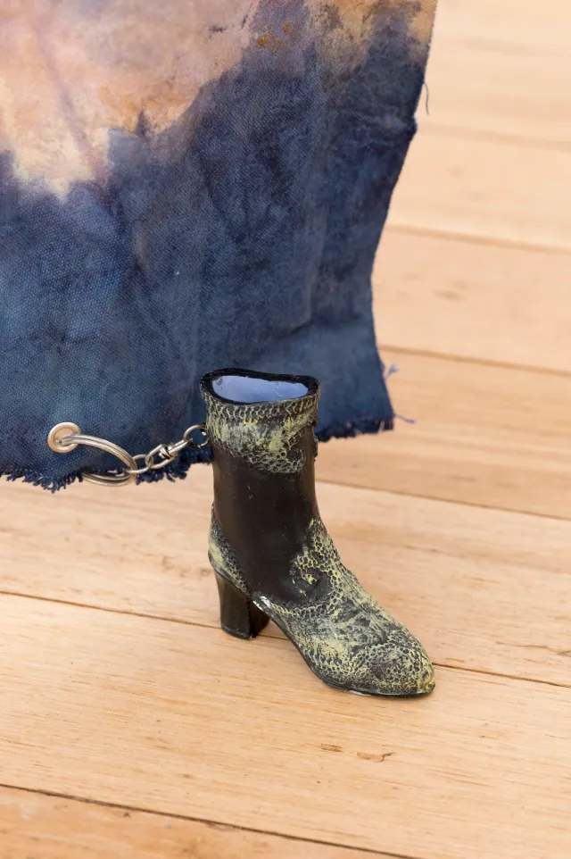 A black heeled boot with worn cream paint across the ankle and toe sits on light-coloured floorboards. The inside of the boot is light blue. The back of the boot is chained to a metal ring which is attached to a hanging piece of worn blue fabric. The fabric is discoloured along the top in an even section.