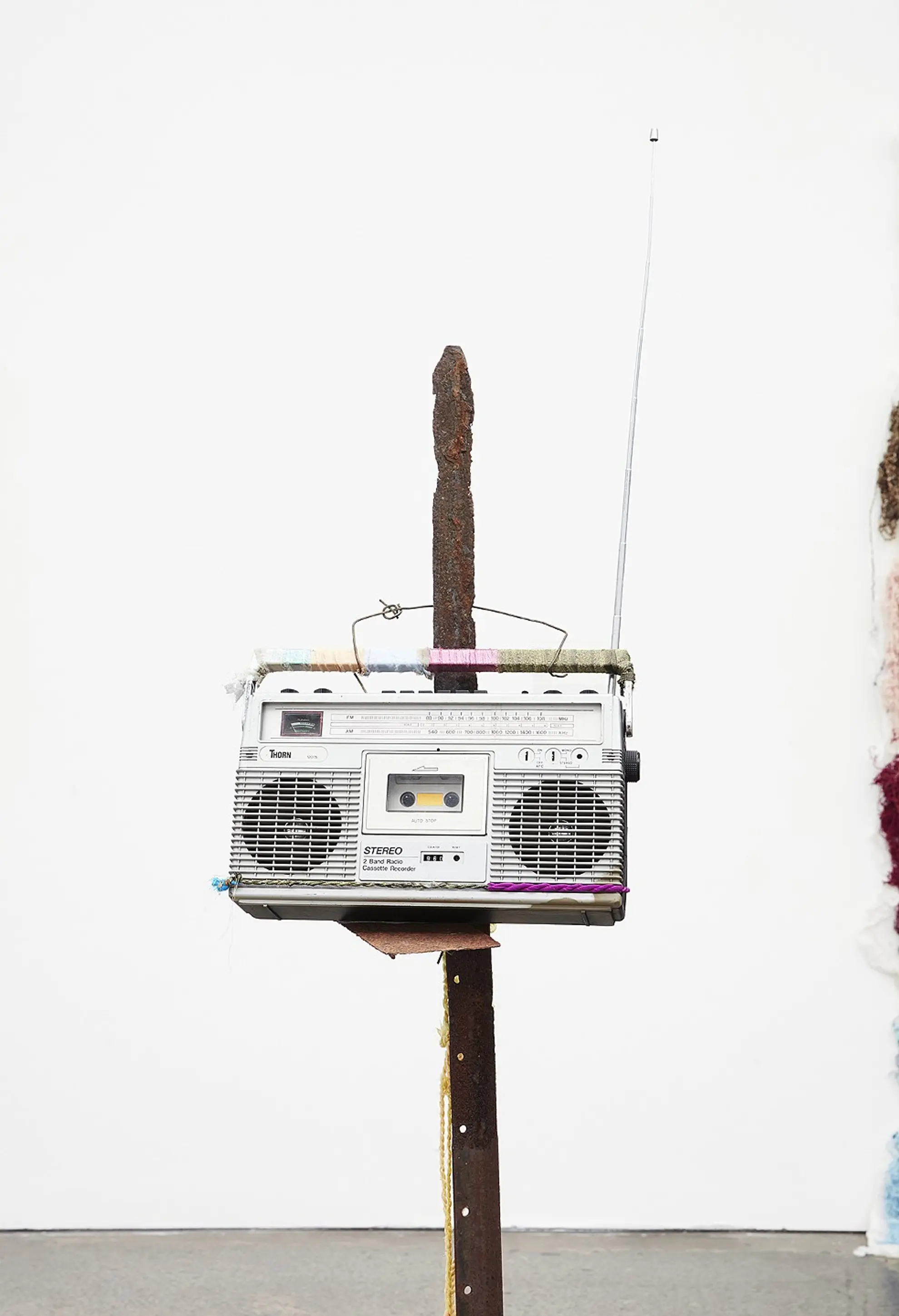 An old rectangular, silver stereo hangs on the end of an upright, rusted fence pole with a point. The stereo has a tall antenna.